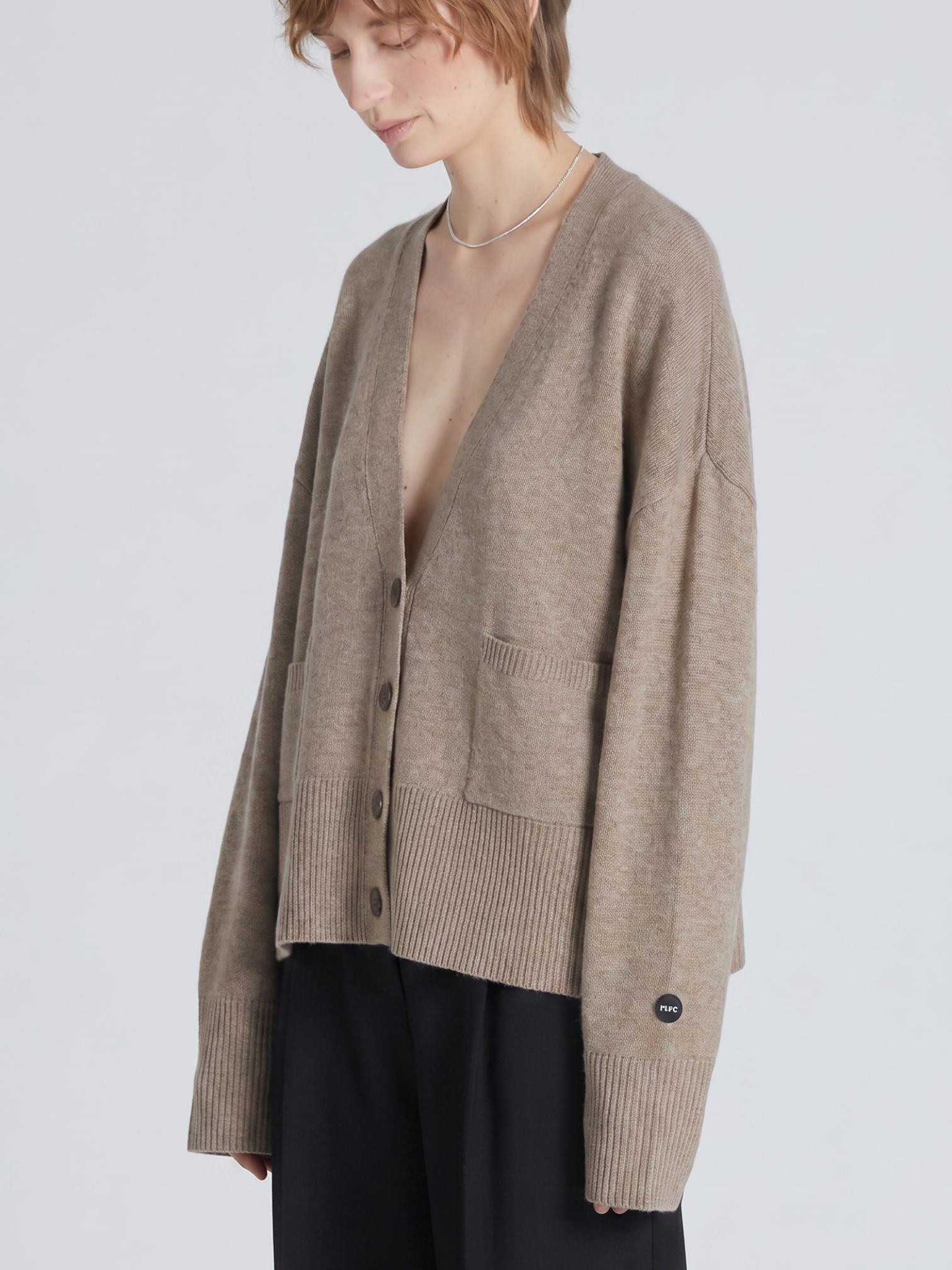 A model wearing a taupe knit cardigan with dark buttons, paired with black wide-leg trousers, against a light gray studio background.