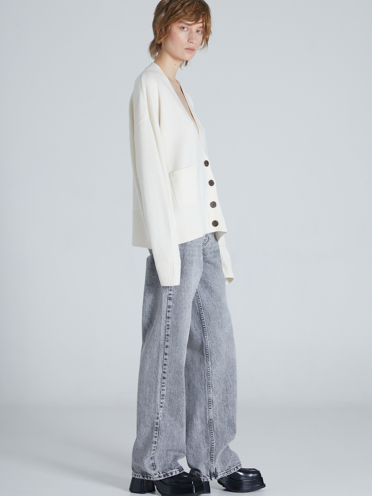 A model is shown from a side profile wearing an oversized off-white cardigan with dark buttons, wide-leg light gray jeans, and black platform shoes, set against a plain light gray studio background.