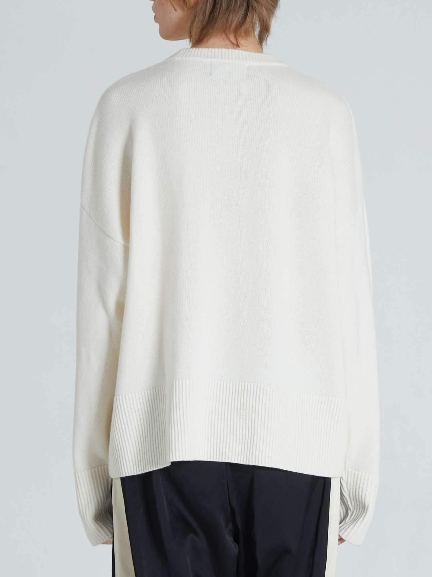 A person shown from the back wearing an oversized cream knit sweater and black pants against a light gray studio background.