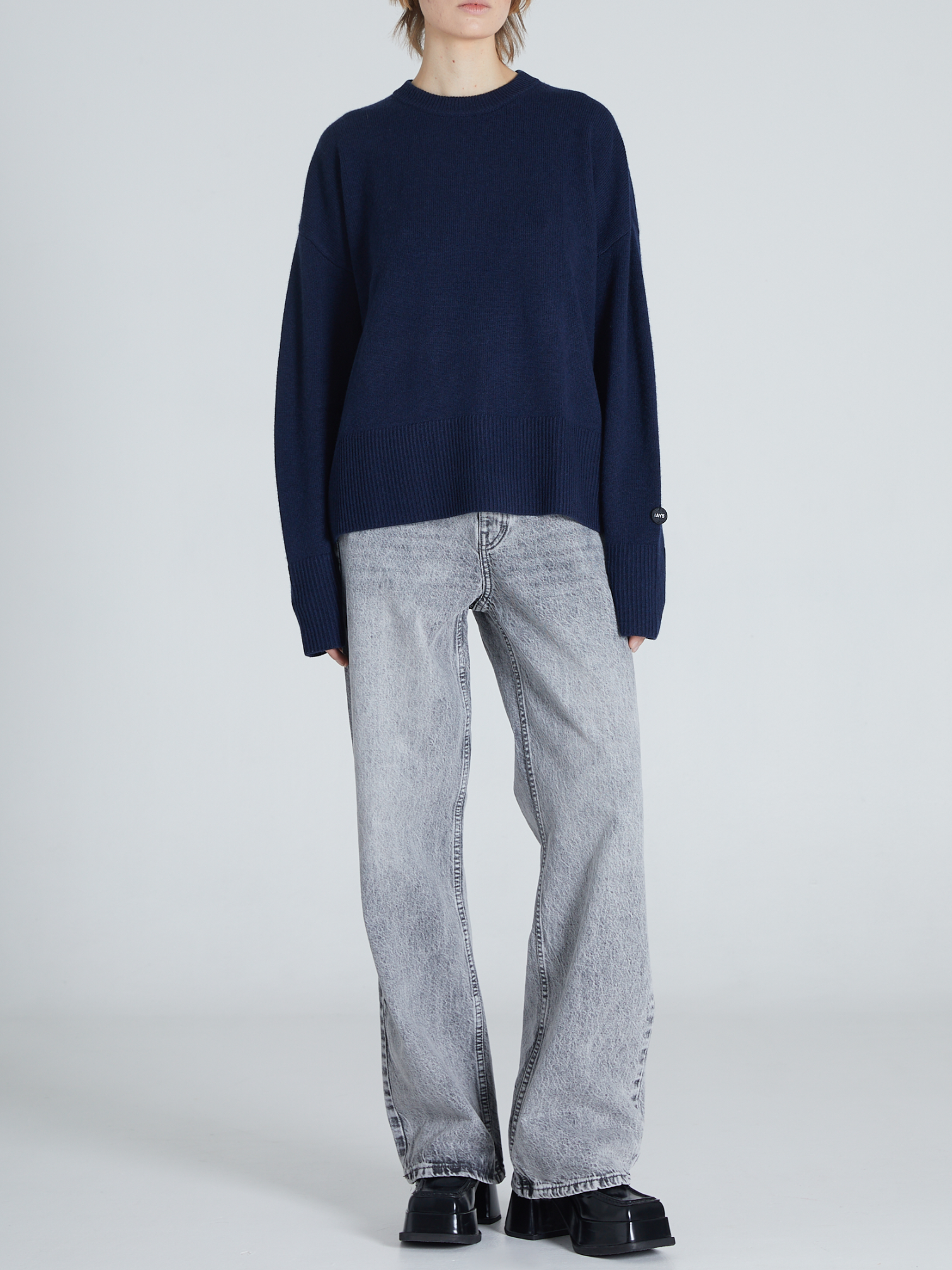 A model wearing a dark navy blue knit sweater and light gray wide-leg jeans, standing against a light gray studio background.