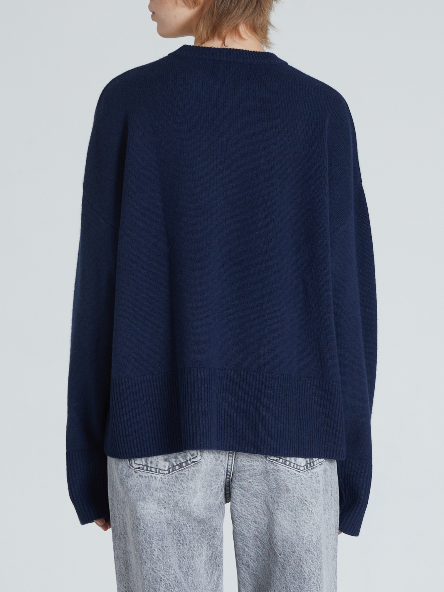 A person shown from the back wearing an oversized navy blue knit sweater and light gray jeans against a plain light gray studio background.