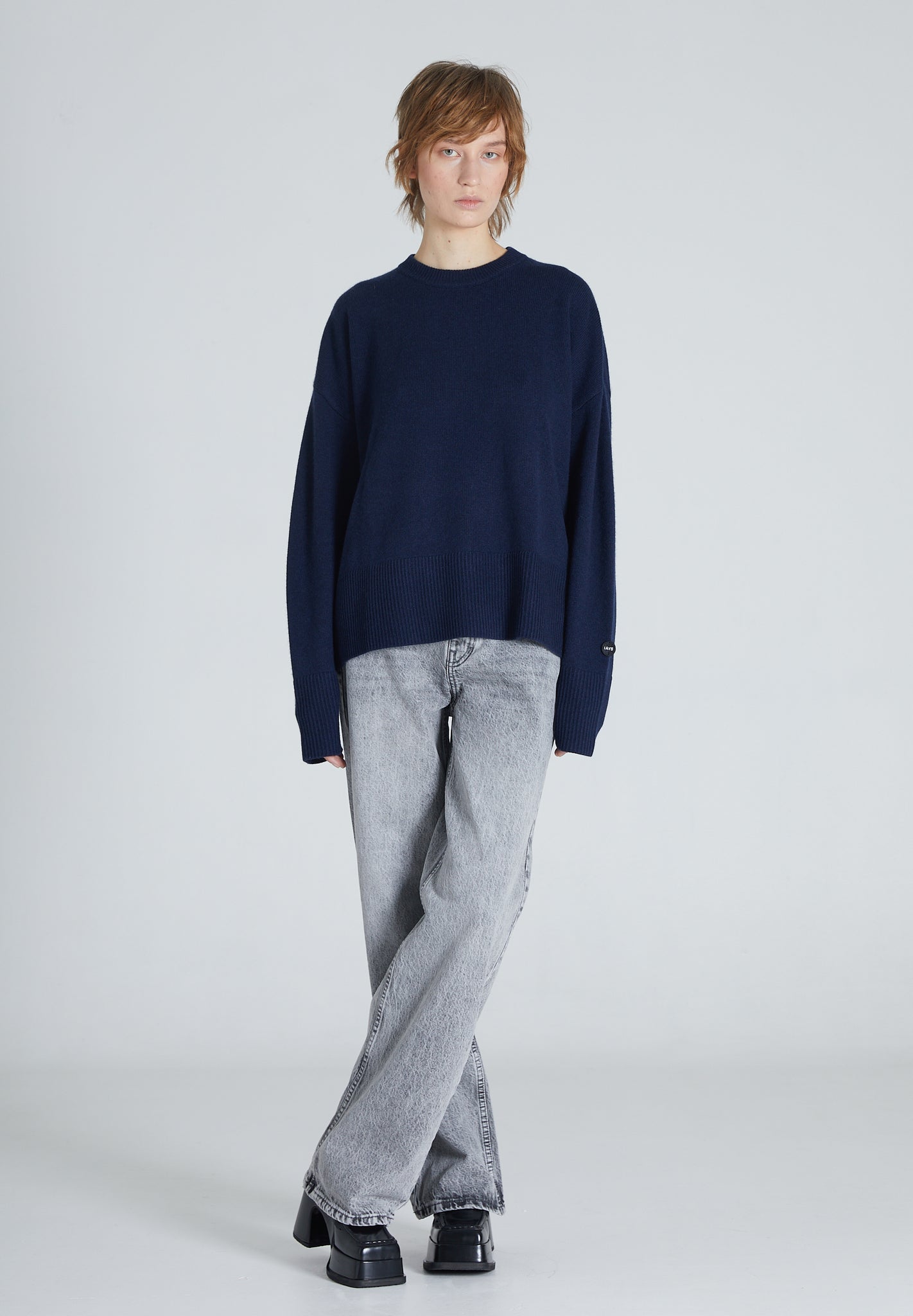 A woman wearing a navy cashmere crew neck sweater and blue jeans, standing against a plain background.