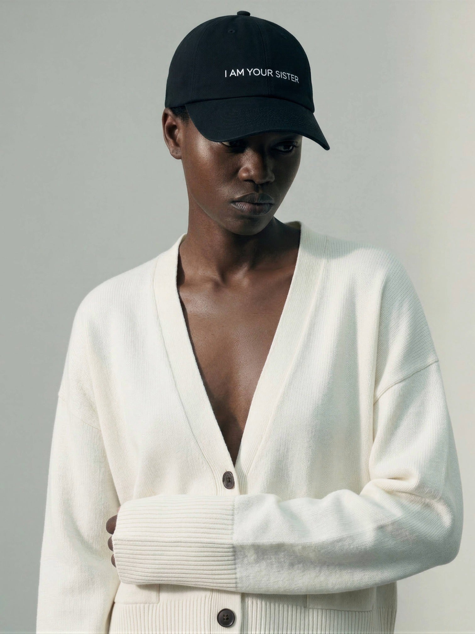 Woman wearing a black cap with 'I AM YOUR SISTER' text and a white cardigan on a gray background