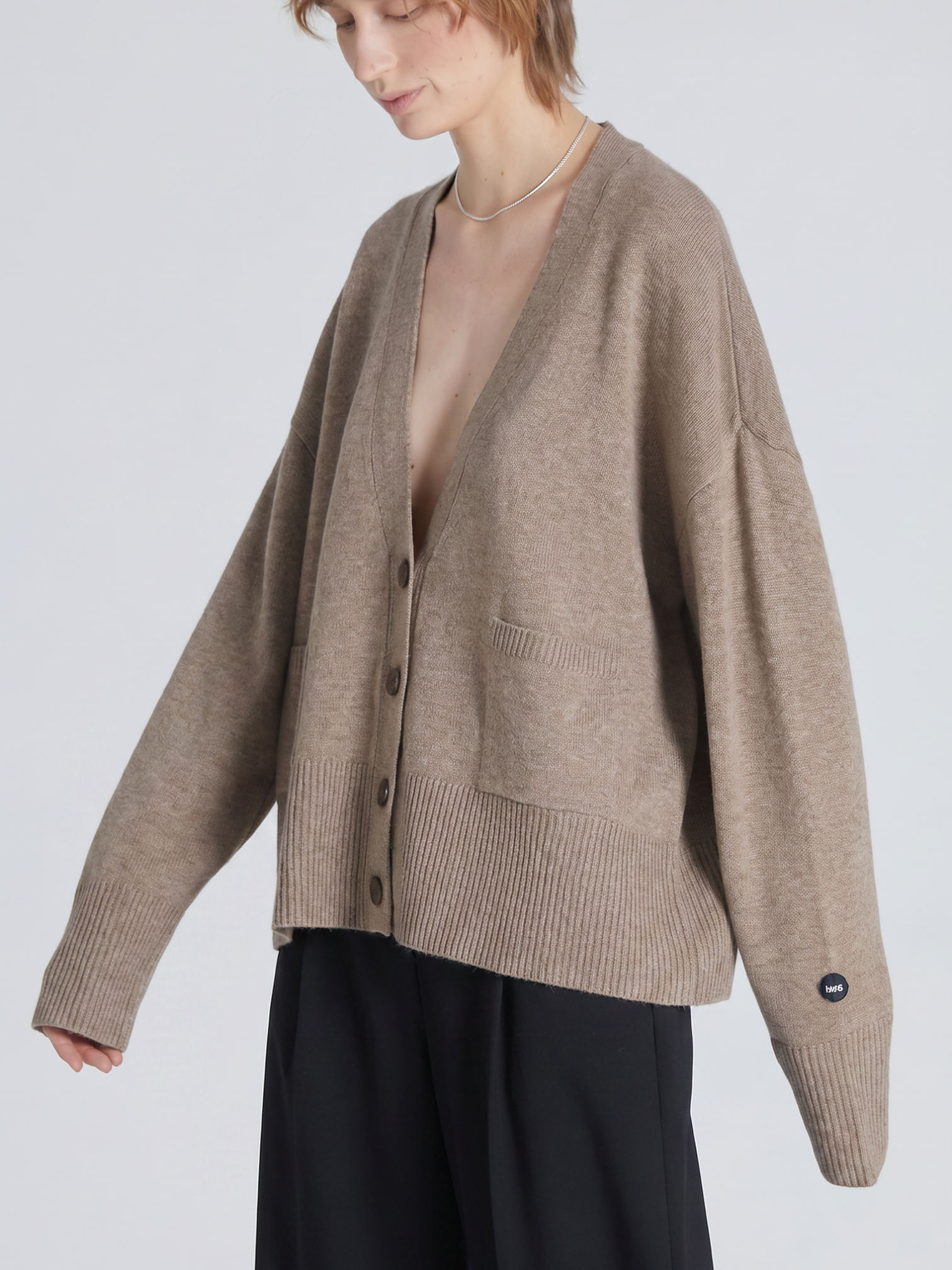 A model wearing an oversized taupe knit cardigan with dark buttons, ribbed trim, and a small black circular sleeve tag, paired with black wide-leg trousers against a light gray studio background.