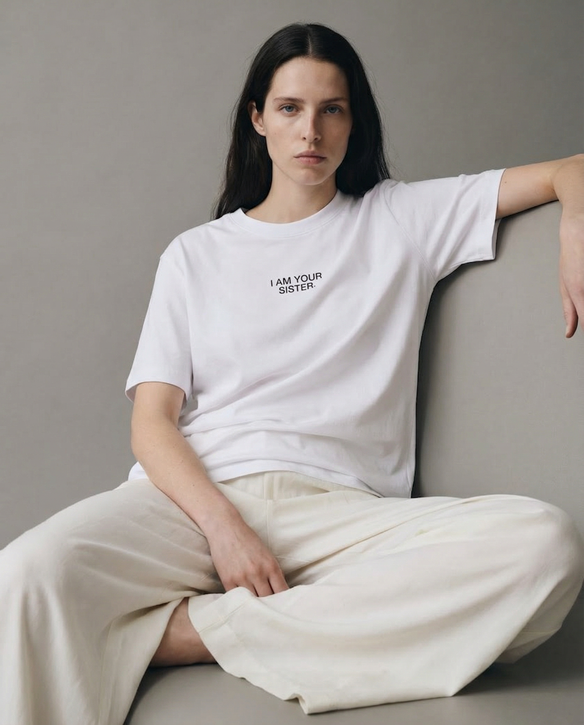 Woman wearing a white I am your sister t-shirt, sitting on a gray couch against a gray background