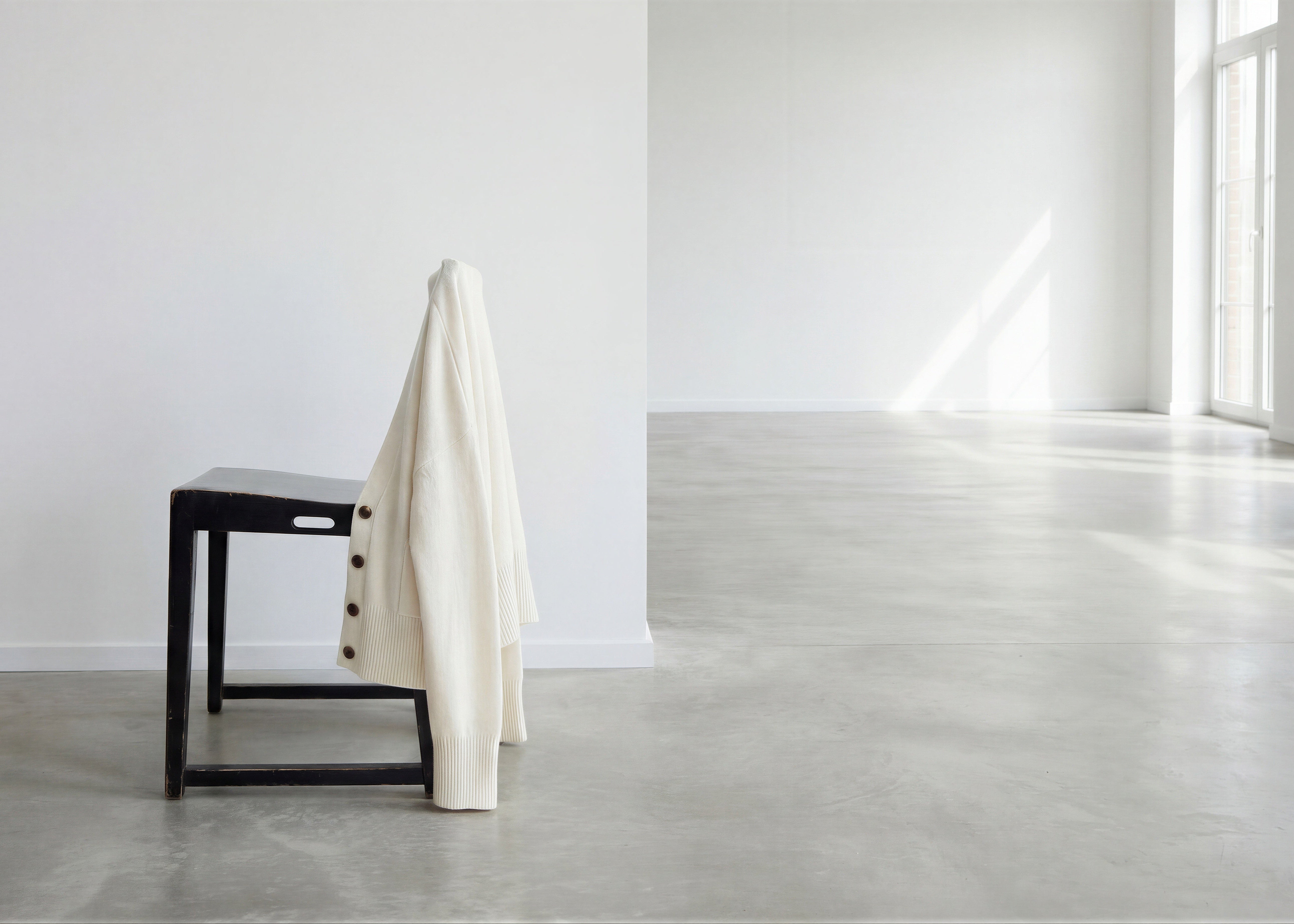 White coat draped over a black chair in a minimalistic room with concrete floor and large windows.