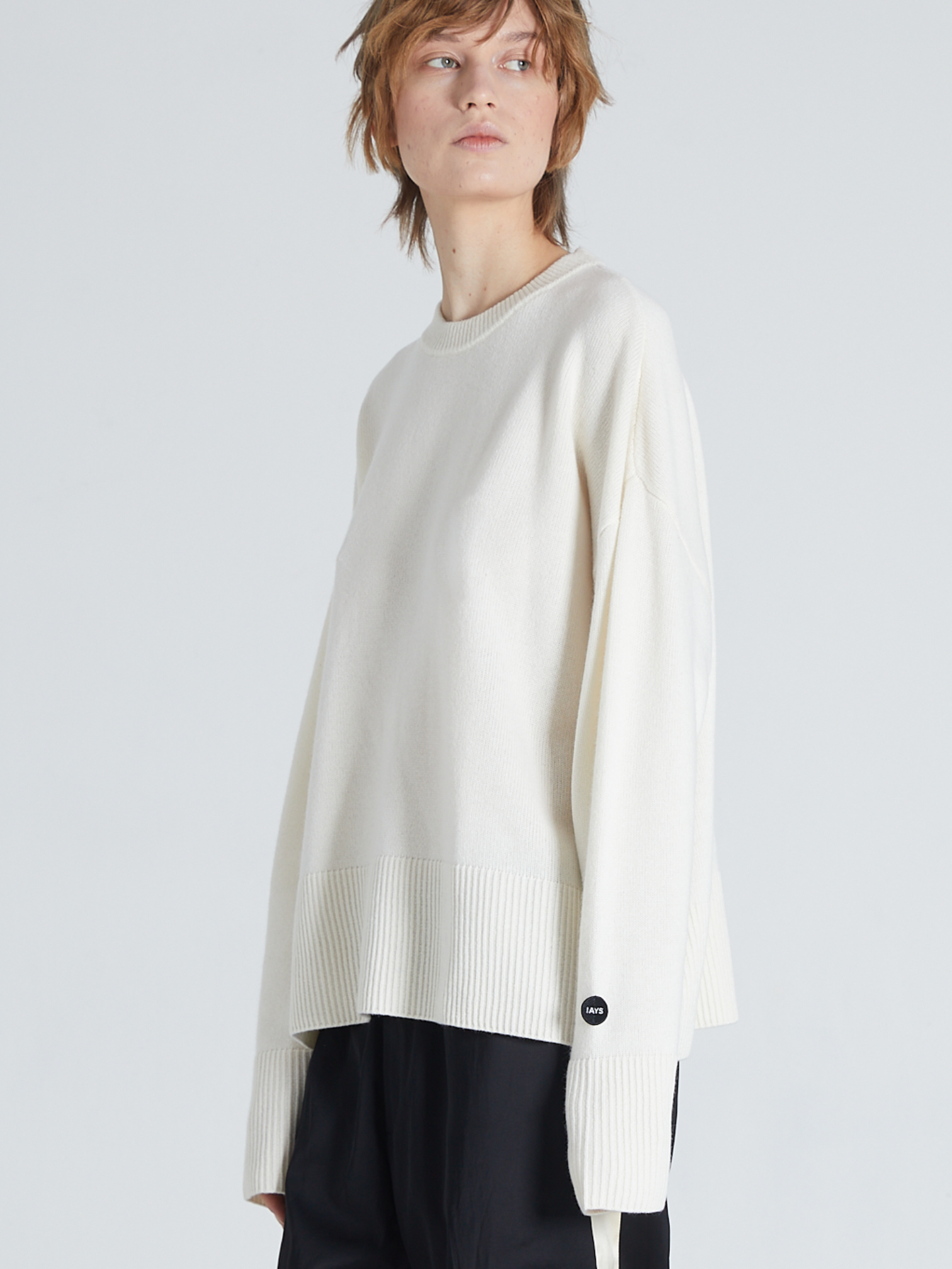 A model wearing an oversized off-white knit sweater with long sleeves and a ribbed hem, paired with black trousers, against a light gray studio background.