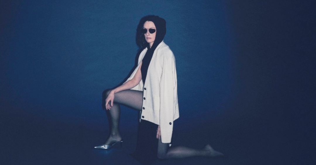 A model wearing a white button-up jacket, black top, black skirt, black tights, and silver pointed heels, kneeling on a dark blue studio background.