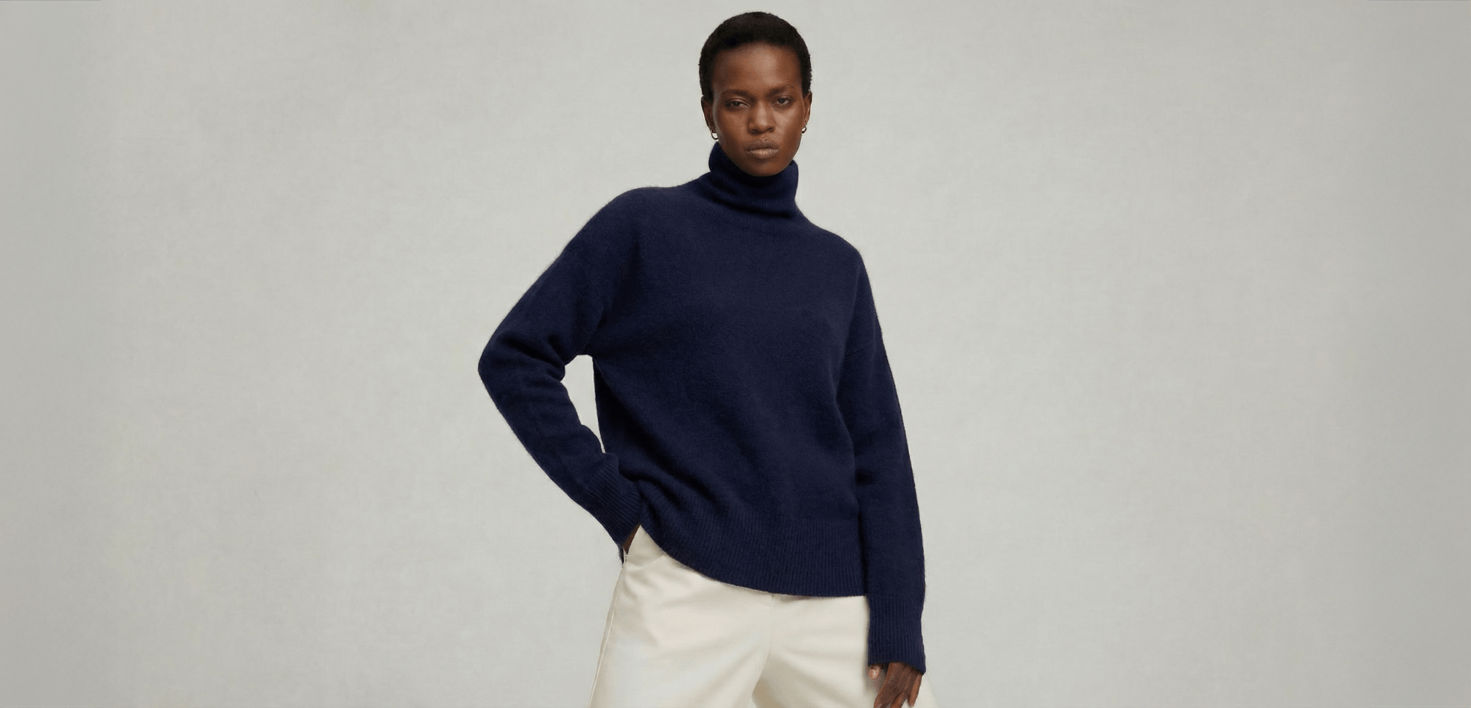 Woman is wearing a navy blue turtleneck cashmere sweater and white pants on a gray background