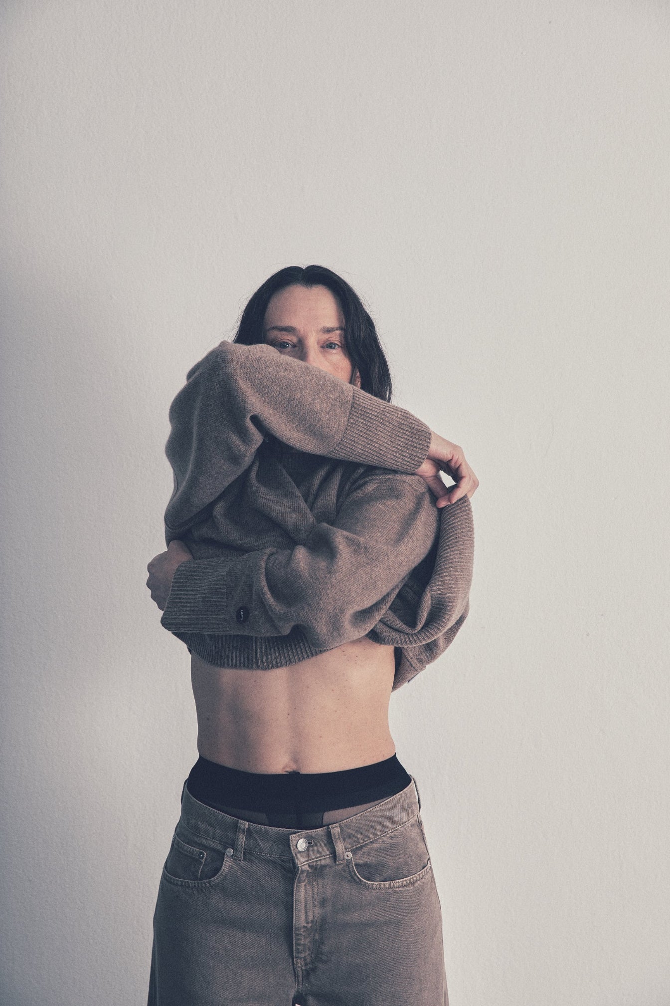 A woman wearing a brown knit cardigan sweater and gray jeans, holding the cardigan up over her head to show its fit and texture against a plain light gray wall.
