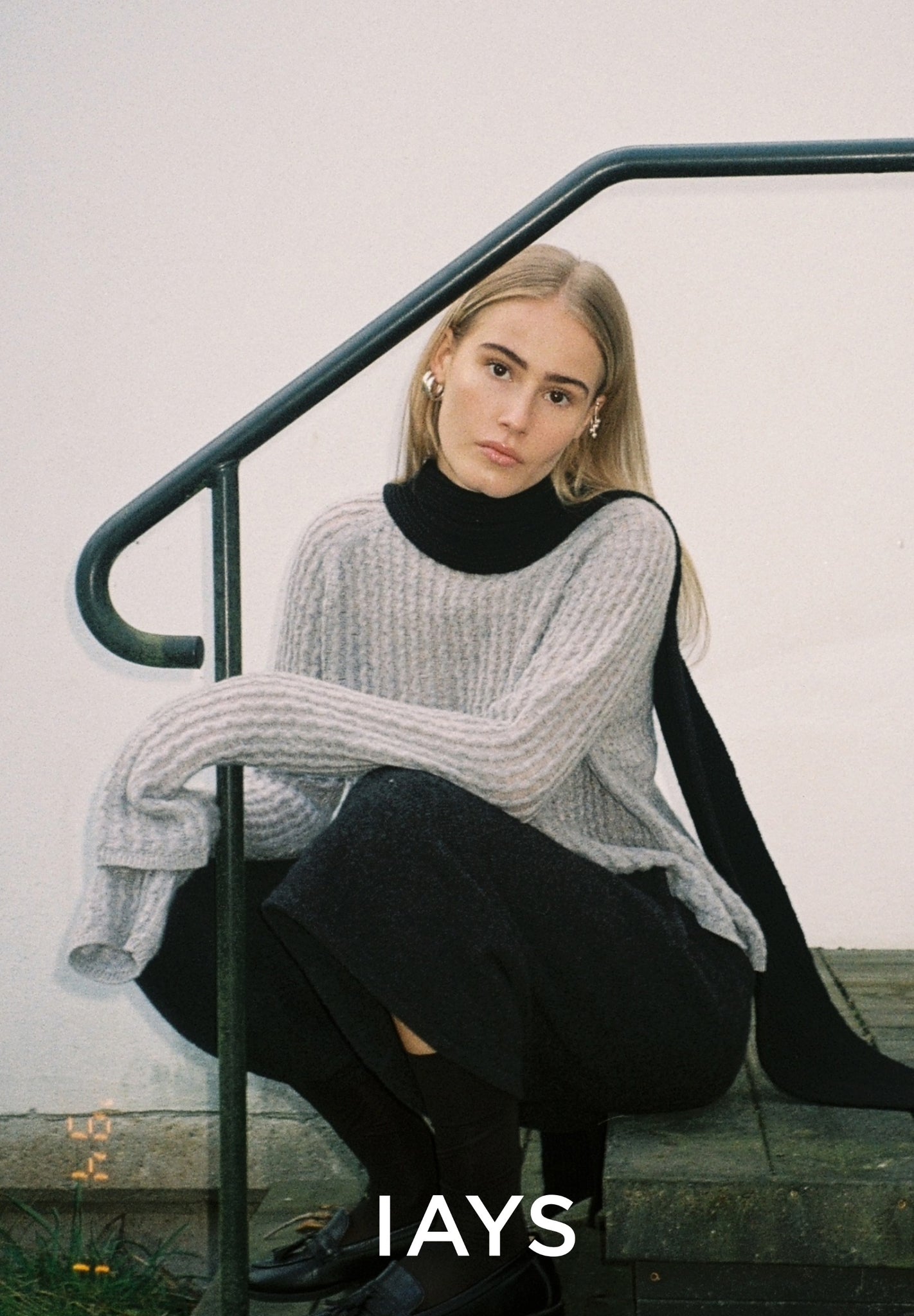 A woman wearing a gray sweater and black pants is sitting on a metal railing with 'IAYS' branding.