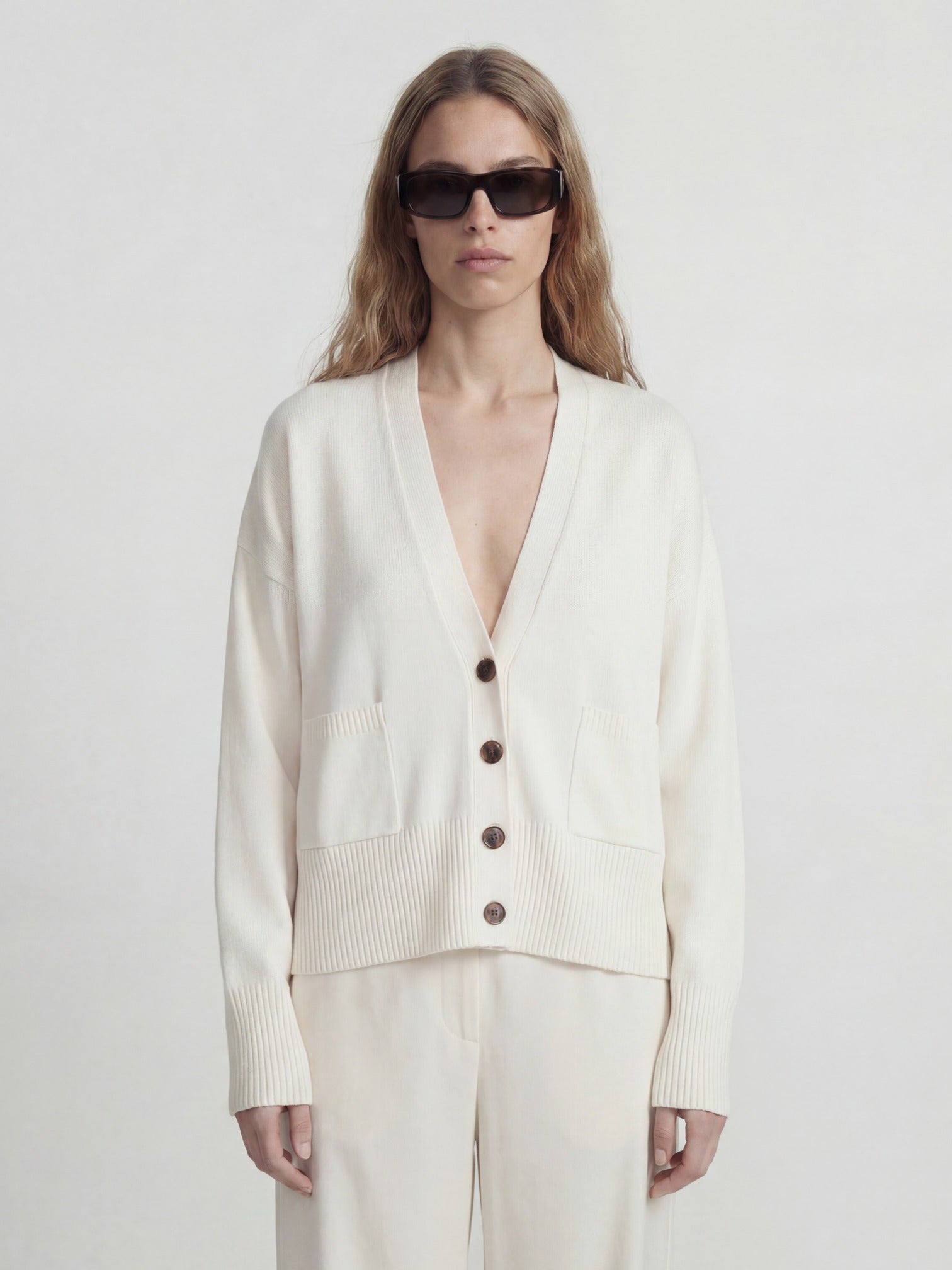 Woman wearing a white cashmere cardigan with buttons and light-colored pants on a plain background