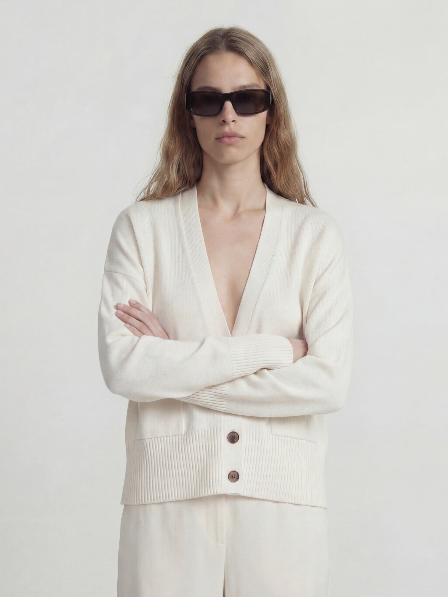 woman wearing a cream-colored cashmere cardigan over a white top with sunglasses on a plain background