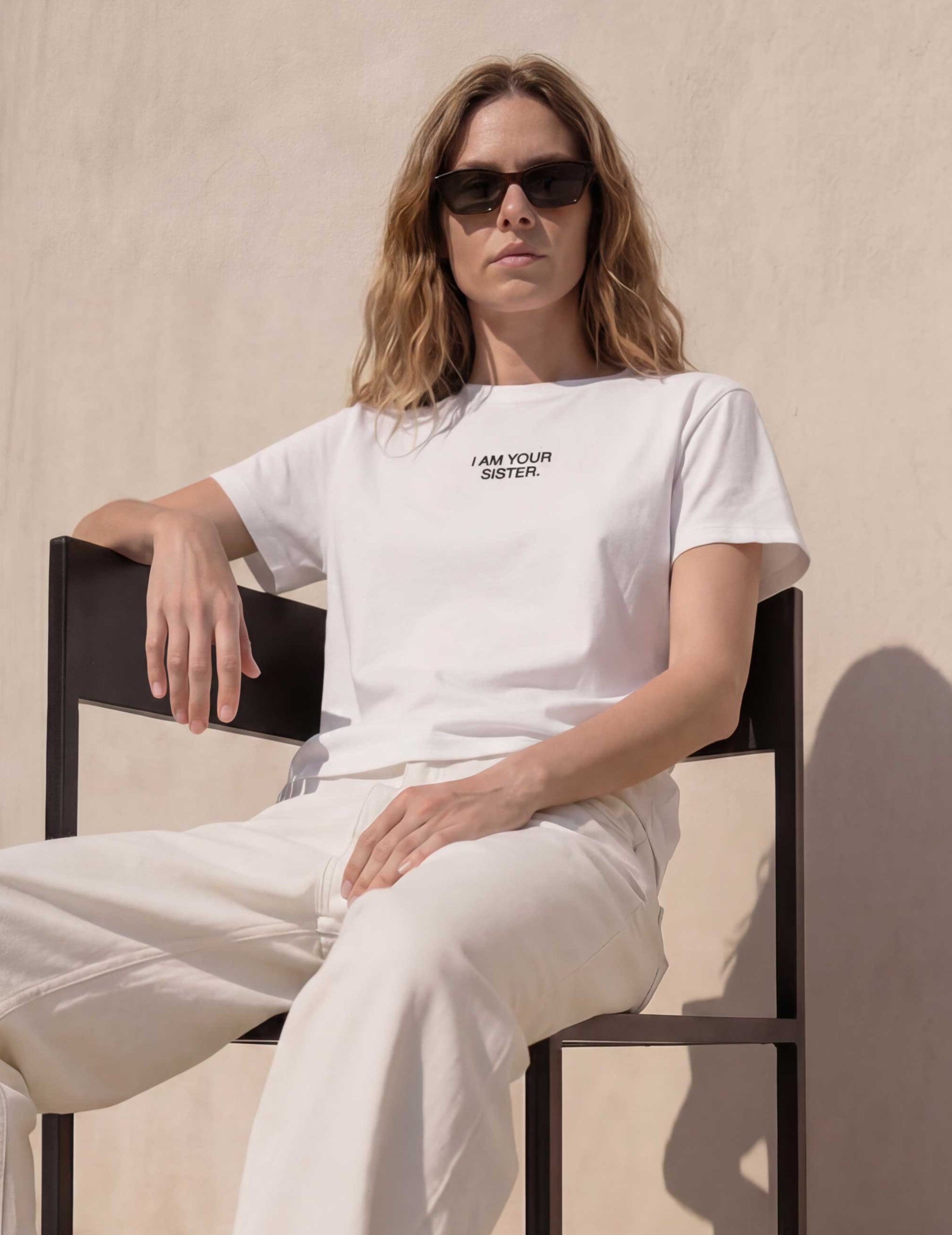Woman wearing a white I am your sister t-shirt and pants sitting on a chair against a beige wall.
