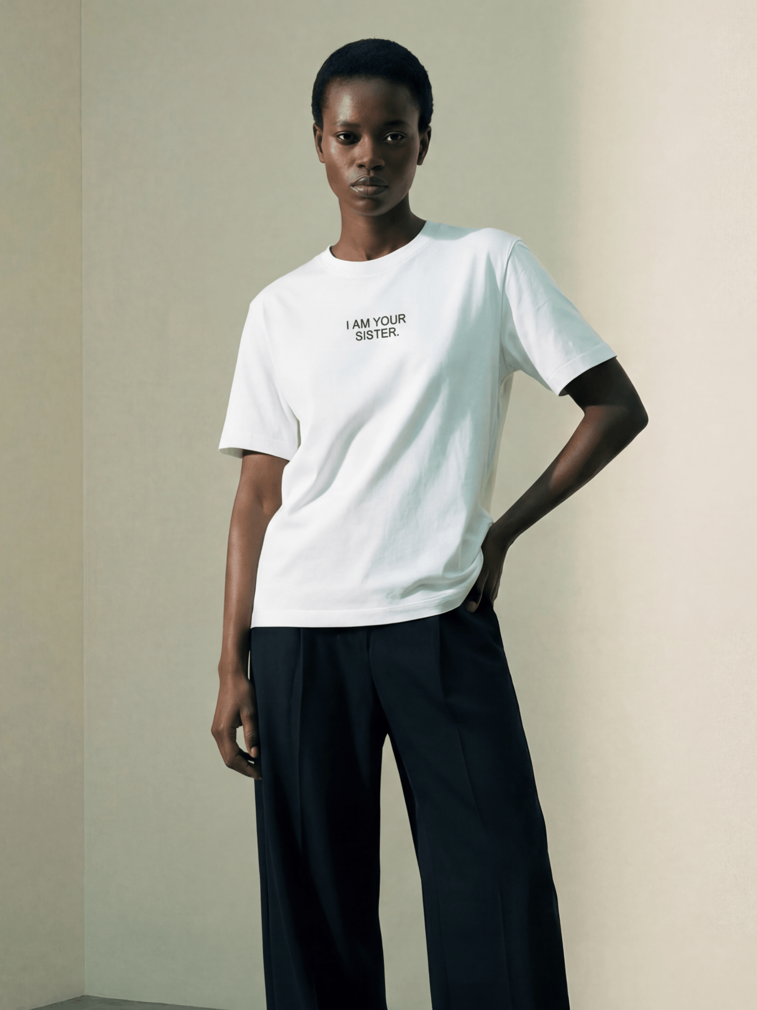 Woman wearing a white Sister Tee with text, standing against a plain background