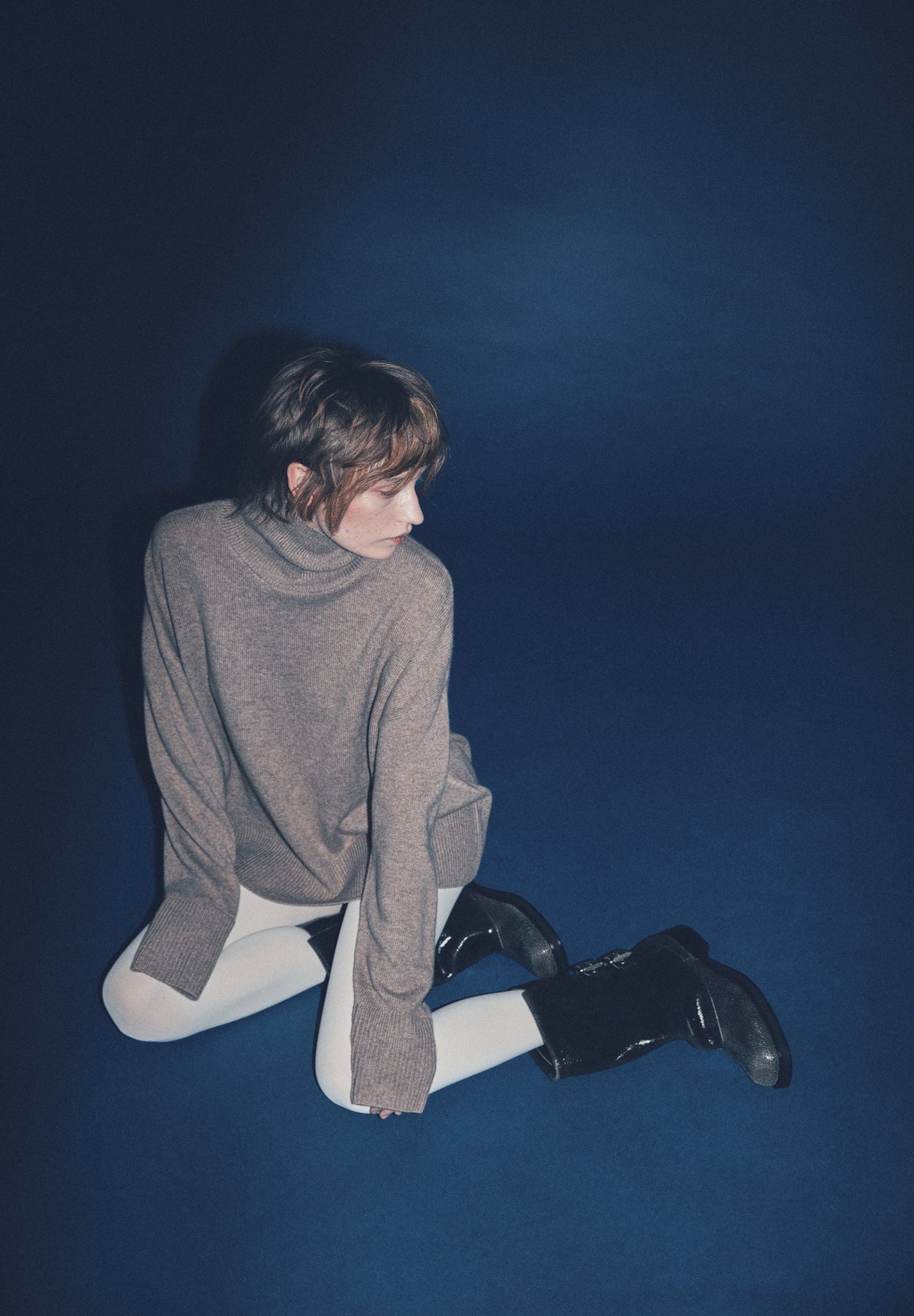 A woman wearing a brown cashmere turtleneck sweater with ribbed trims, featuring a standing neck, dropped shoulders, and extended sleeves, is sitting against a blue background.