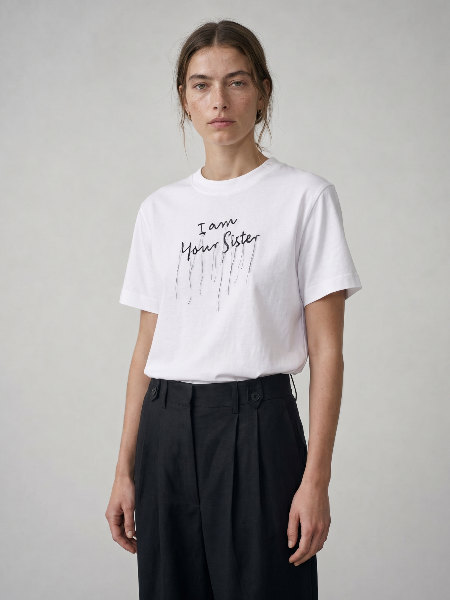 A model wearing a white short-sleeve t-shirt with 'I am Your Sister' embroidery in black script with a dripping paint effect, paired with black trousers, against a neutral gray background.
