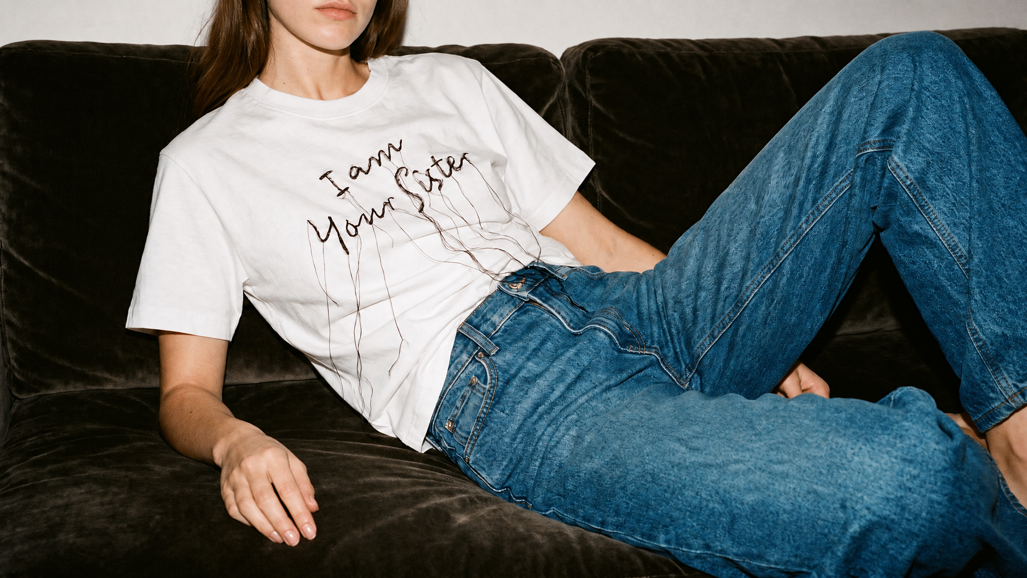 A woman reclining on a dark brown velvet couch, wearing a white graphic t-shirt with the text 'I am Your Sister' in black script with a dripping ink effect, paired with blue jeans.