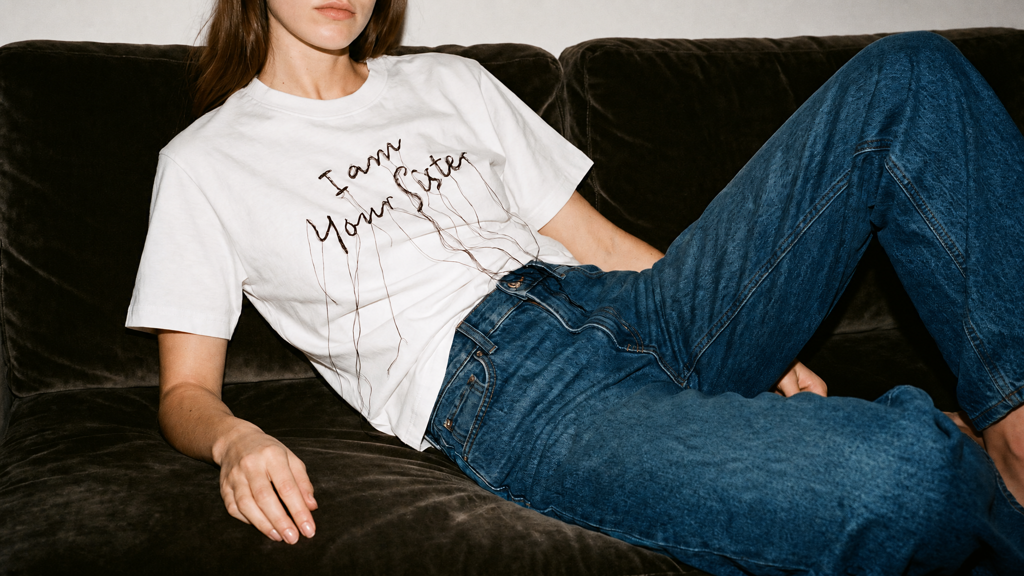 A woman reclining on a dark brown velvet couch, wearing a white graphic t-shirt with the text 'I am your sister' in black script with a dripping ink effect, paired with blue jeans.